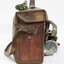 Proper right side of a of a Victorian Railways guard's satchel, with a rolled up green signal flag attached behind the handle on the top and a dark blue metal lamp attached to the front .