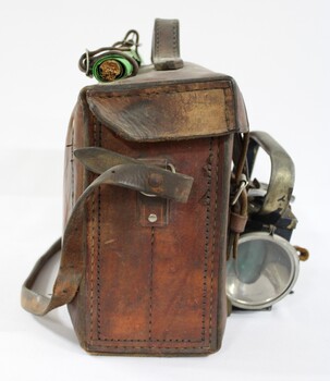 Proper right side of a of a Victorian Railways guard's satchel, with a rolled up green signal flag attached behind the handle on the top and a dark blue metal lamp attached to the front .