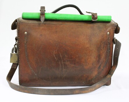 Back view of a Victorian Railways guard's satchel, with a rolled up green signal flag attached to the top amd a brass padlock on the proper left side.