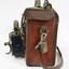 Proper left side of a of a Victorian Railways guard's satchel, with a rolled up green signal flag attached behind the handle on the top, a brass padlock attached to the hardware for the should strap on the side, and a dark blue metal lamp attached to the front.