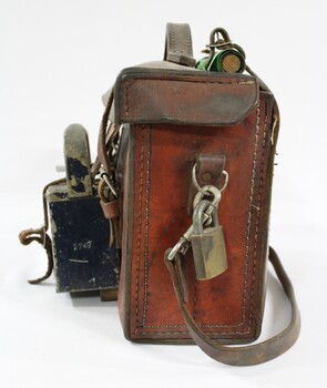 Proper left side of a of a Victorian Railways guard's satchel, with a rolled up green signal flag attached behind the handle on the top, a brass padlock attached to the hardware for the should strap on the side, and a dark blue metal lamp attached to the front.