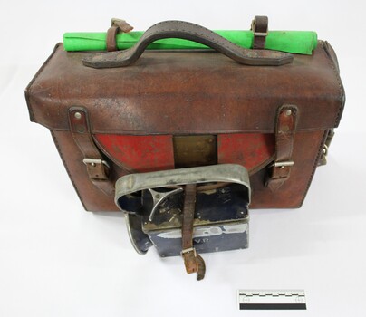 Front and top surface of a Victorian Railways guard's satchel, with a rolled up green signal flag attached behind the handle and a dark blue metal lamp attached to the front, and a black and white 10 cm scale in the foreground..