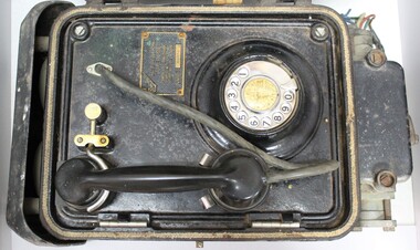 Lower part of a metal case containing a black rotary dial flameproof telephone, with rubber tubing attached to the handset and a metal plate with the maker's name and the type and certification of the telephone.