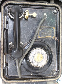 Vertical view of a black rotary dial flameproof telephone, with rubber tubing attached to the handset and a metal plate with the maker's name and the type and certification of the telephone.