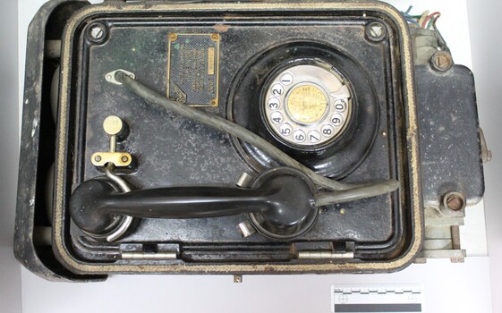 Lower part of a metal case containing a black rotary dial flameproof telephone, with rubber tubing attached to the handset and a metal plate with the maker's name and the type and certification of the telephone, with a black and white 10 cm scale.