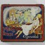 A tobacco tin with a coloured illustration of a woman in stylish 1920s-1930s dress sitting in an armchair smoking a cigarette, while watching an Orientalist scene featuring a belly dancer and a snake charmer wearing a turban. 