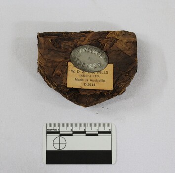 Lump of tobacco with Havelock Tobacco makers mark on tin insert, with manufacture details on paper, pictured with 5cm scale