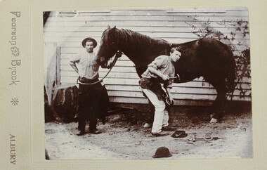 Black and white photograph of two men in front of a weatherboard building. The man on the left is holding onto the bridle while the man on the right is shoeing the horse. 