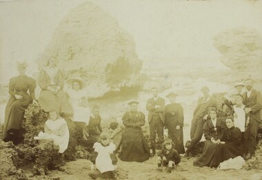 A sepia group portrait of adults and children on a beach, wearing Victorian era clothing. 