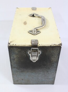 One end of a rectangular grey metal railways lunch box with a cream coloured lid secured with metal clips and a metal handle on the lid.