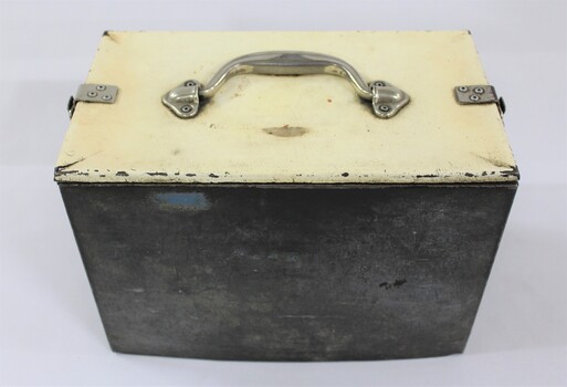 Side view of a rectangular grey metal railways lunch box with a cream coloured lid secured with metal clips on both ends and a metal handle on the lid.