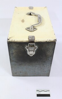 One end of a rectangular grey metal railways lunch box with a cream coloured lid secured with metal clips and a metal handle on the lid, with a black and white 5 cm scale.