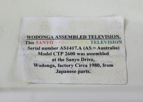 Paper label attached to the upper back part of the Sanyo VHF Colour Television with details about its manufacture, which is discolured and peeling  at the corners.