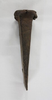 Side view of a hand forged iron alloy wedge with a cracked flange/rim at the top, red-brown rust and some dirt on the surface.