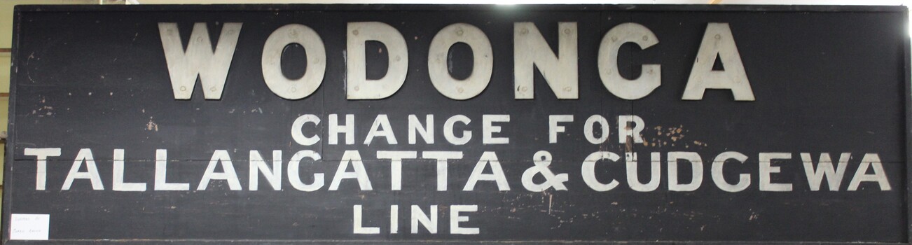 Painted wooden sign from the Wodonga train station with the directions for changing to the Tallangatta and Cudgewa train line in light coloured paint on a black background.