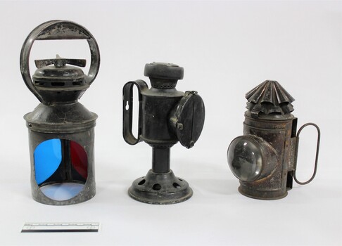 Three metal and glass lamps, one with blue and red filters, used for signalling on the Victorian Railways, with a black and white 10 cm scale to indicate their size.