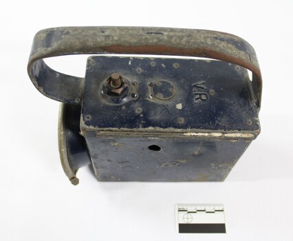 View from above of a rectangular dark blue painted metal lamp with a large handle on the top and 'V.R.' for Victorian Railways embossed on the top and proper left side andsurfaces, and a black and white 5 cm scale.