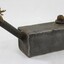 Proper let side of a rectangular grey metal lamp with a textile fibre wick protruding from a long spout at one end, a flat handle at the other end and a cork inserted in the opening on the top surface.