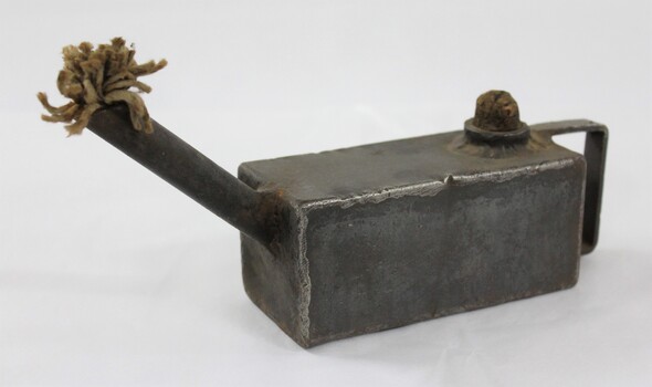 Proper let side of a rectangular grey metal lamp with a textile fibre wick protruding from a long spout at one end, a flat handle at the other end and a cork inserted in the opening on the top surface.