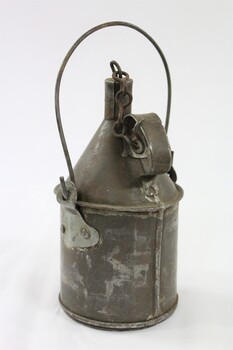 Back of a grey metal container with a looped handle attached to the sides, as well as a handle on the back of the container with a metal chain attached to it to secure the cap for the container. 