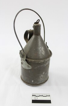 Grey metal container with a looped handle attached to the sides, as well as a handle on the back of the container with a metal chain attached to it to secure the cap for the container, and a black and white 5 cm scale in the foreground.