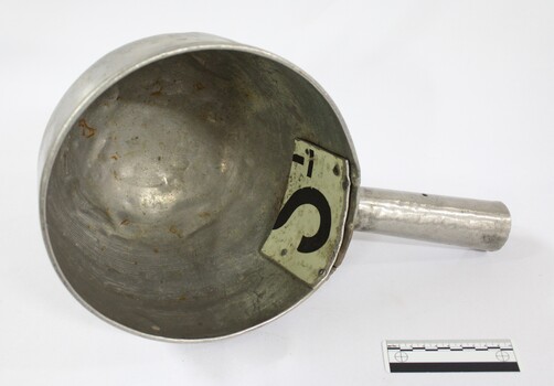 Interior of a large aluminium scoop with a handle on one side edge and a repair riveted to the interior surface where the handle is soldered onto the bowl of the scoop. Black and white 10 cm scale in the foreground.