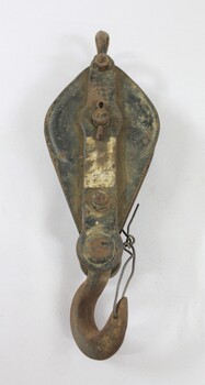 Front of an elongated ellipse-shaped metal pulley, painted black with a lot light brown dirt on the surface and traces of a silver coloured rectangular label on the central raised section.