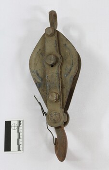 Back of an elongated ellipse-shaped metal pulley, painted black with a lot light brown dirt on the surface and a black and white 5 cm scale to one side of the pulley.