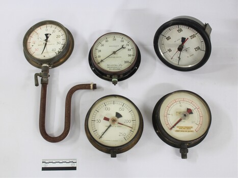Five metal and glass water pump pressure gauges, including one with a U-shaped pipe and lever attached, with a black and white 10 cm in the foreground.