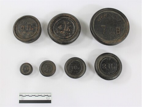 Set of seven metal weights with imperial measurements from 4 oz. to 7 lb., with a black and white 10 cm scale in the foreground.