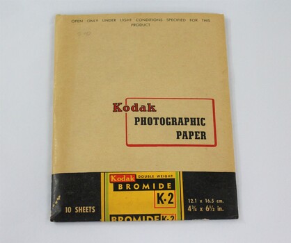 A paper packet coloured light tan, black green, yellow and red, containing Kodak photographic paper.