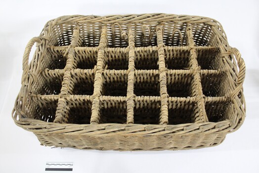 Rectangular wicker crate for delivering bottled drinks with 12 compartments and handles at each narrow end, and a black and white 10 cm scale in the foreground.
