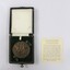 Circular bronze medal depicting a man and a woman riding a horse, in a small presentation box with a dark green velvet insert for the medal in the lower part and a light coloured textile printed with the dates 1851 and 1951 in the lid. Text on paper with a description of the medal next to the case.