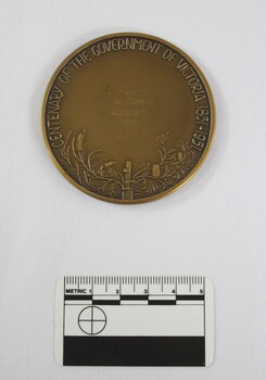 Reverse of a circular bronze medal with a stake surrounded by Australian floral motifs in the lower part and the words "CENTENARY OF THE GOVERNMENT OF VICTORIA 1851-1951" in raised letters around the top edge, and "Preseneted to / The Shire of / Wodonga / 1951" in engraved letters in the centre.  Black and white 5 cm scale in the foreground.