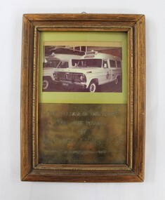 Wooden frame around a metal plaque commemorating the purchase of an ambulance made posible by the Wodonga Lions Club in August 1971. Photograph of two ambulances included in the frame above the metal plaque.