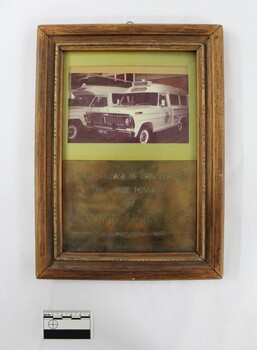 Wooden frame around a metal plaque commemorating the purchase of an ambulance made posible by the Wodonga Lions Club in August 1971. Photograph of two ambulances included in the frame above the metal plaque. Black and white 5 cm scale in the foreground.