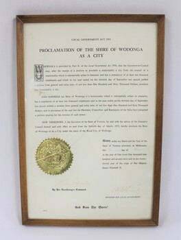 Thin wooden frame on a certificate with the text in black proclaiming the Shire of Wodonga as a City dated to 1973. Embossed gold seal on the left side of the text in the  lower part of the certificate.