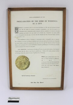 Thin wooden frame on a certificate with the text in black proclaiming the Shire of Wodonga as a City dated to 1973. Embossed gold seal on the left side of the text in the  lower part of the certificate. Black and white 5 cm scale in the foreground.