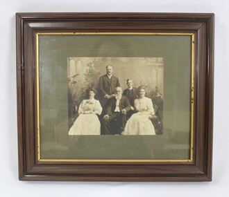 Sepia-toned photograph of John Woodland and his family in a brown wooden frame with a gold bevelled border and grey matt board around the photo. 
