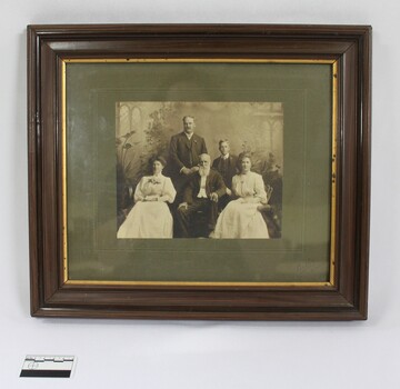 Sepia-toned photograph of John Woodland and his family in a brown wooden frame with a gold bevelled edge and grey matt board around the photo. Black and white 5 cm scale in the foreground.