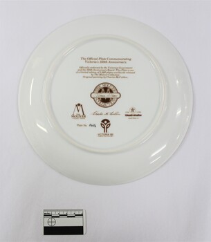 Back of a ceramic plate commemorating the 150th anniversary of Victoria, with an inscription about the plate and the artist's signature, as well as the plate number 0404 and three trade marks. Black and white 5 cm scale in the foreground.