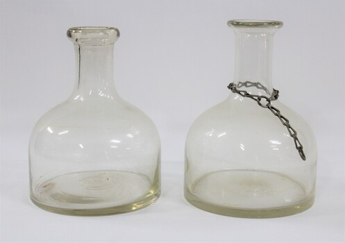 Side view of two clear glass water containers/bottles, one with a metal chain around the narrow neck. 