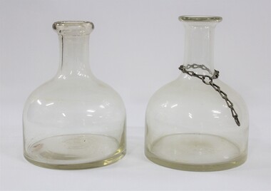 Side view of two clear glass water containers/bottles, one with a metal chain around the narrow neck. 