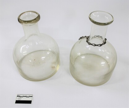 Two clear glass water containers/bottles, one with a metal chain around the narrow neck.  Black and white 5 cm scale in the foreground.