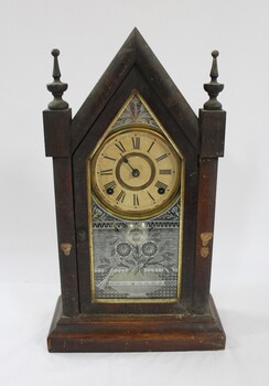 A gothic style wooden clock with gold and white embellishments, and Roman Numeral numbering on the clock face