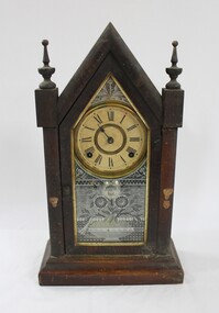 A gothic style wooden clock with gold and white embellishments, and Roman Numeral numbering on the clock face