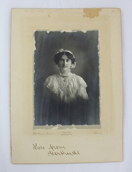 A black and white mounted photograph of a woman in a white, frilled dress 