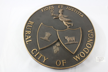 A metal circular plate representing the Rural City of Wodonga. Includes symbols of cattle, wheat and grapes, the mythological figure Hermes/Mercury  and the Latin "Fides et Justitia"