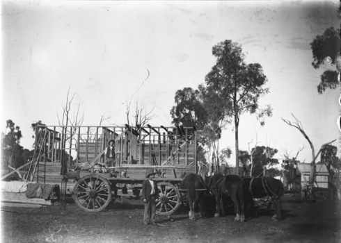 Framework of a house under construction. Two men are working on the site and a group of horses are on the right.