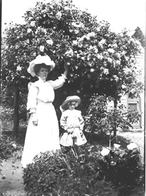  A women and young girl standing under a flowering bush. There is a house in the background.
